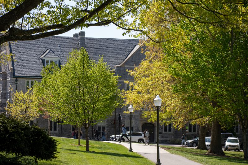 Green trees, grass, and lamp posts surround a pathway leading to grey Hokie Stone buildings in the distance. A few people wearing backpacks walk in the background near parked cars.